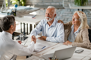 a senior couple meeting with a male financial advisor at a desk where the male client is looking directly at the advisor and shaking the advisor’s hand to symbolize getting started with an advisory relationship.